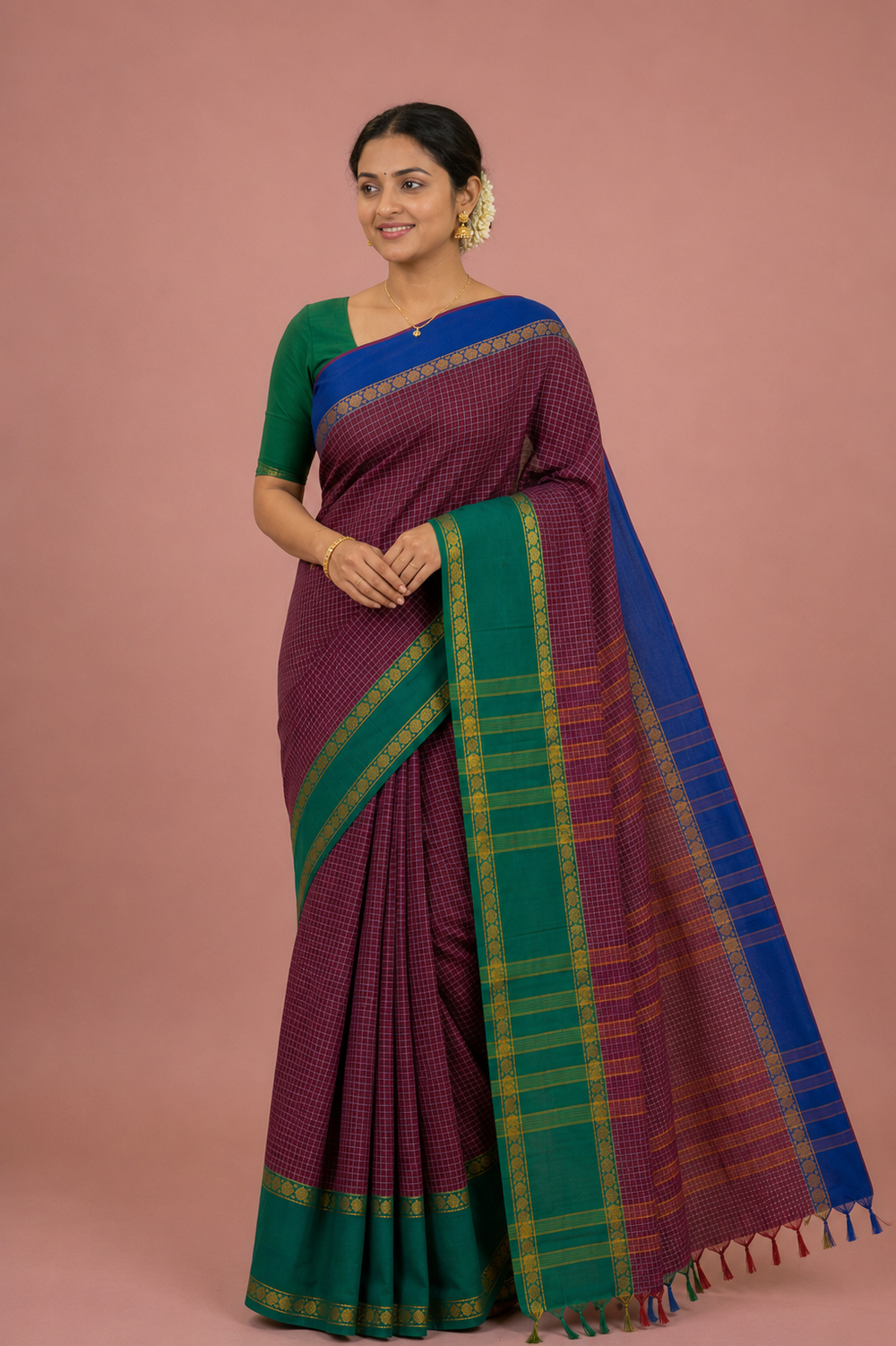 A woman modeling a traditional Indian saree against a solid dusty pink background. The saree features a deep purple body with a small checkered pattern, a green border, and a wide blue pallu with green and yellow stripes.